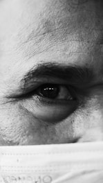 A detailed close-up of an individual's eye and eyebrow is visible, with the skin texture and fine lines accentuated. The lower part of the face is partially covered by a surgical mask, contributing to the foreground elements.
