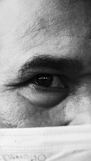 A detailed close-up of an individual's eye and eyebrow is visible, with the skin texture and fine lines accentuated. The lower part of the face is partially covered by a surgical mask, contributing to the foreground elements.