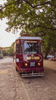 A vintage red tram named 'Pat Rani' is positioned on tram tracks surrounded by lush green trees. The tram has a decorative crown-like structure on its roof, with 'Reserved' sign displayed at the front, and a softly lit interior. The cobblestone path extends into the distance, hinting at a peaceful, charming street setting.