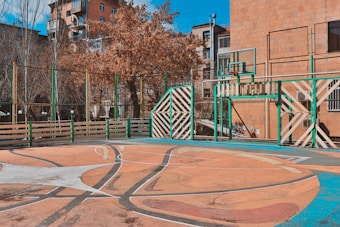 An outdoor basketball court surrounded by a fence with green and white striped metal panels. The court has a well-worn surface with a faded, colorful basketball design. Trees with brown leaves and buildings frame the background, creating an urban setting.