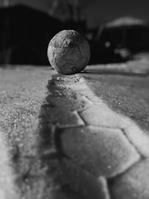 A worn-out soccer ball rests on a textured, uneven surface, casting a long shadow. The background is blurred, emphasizing the ball's position on the ground. The image is in black and white, enhancing the contrast and depth.