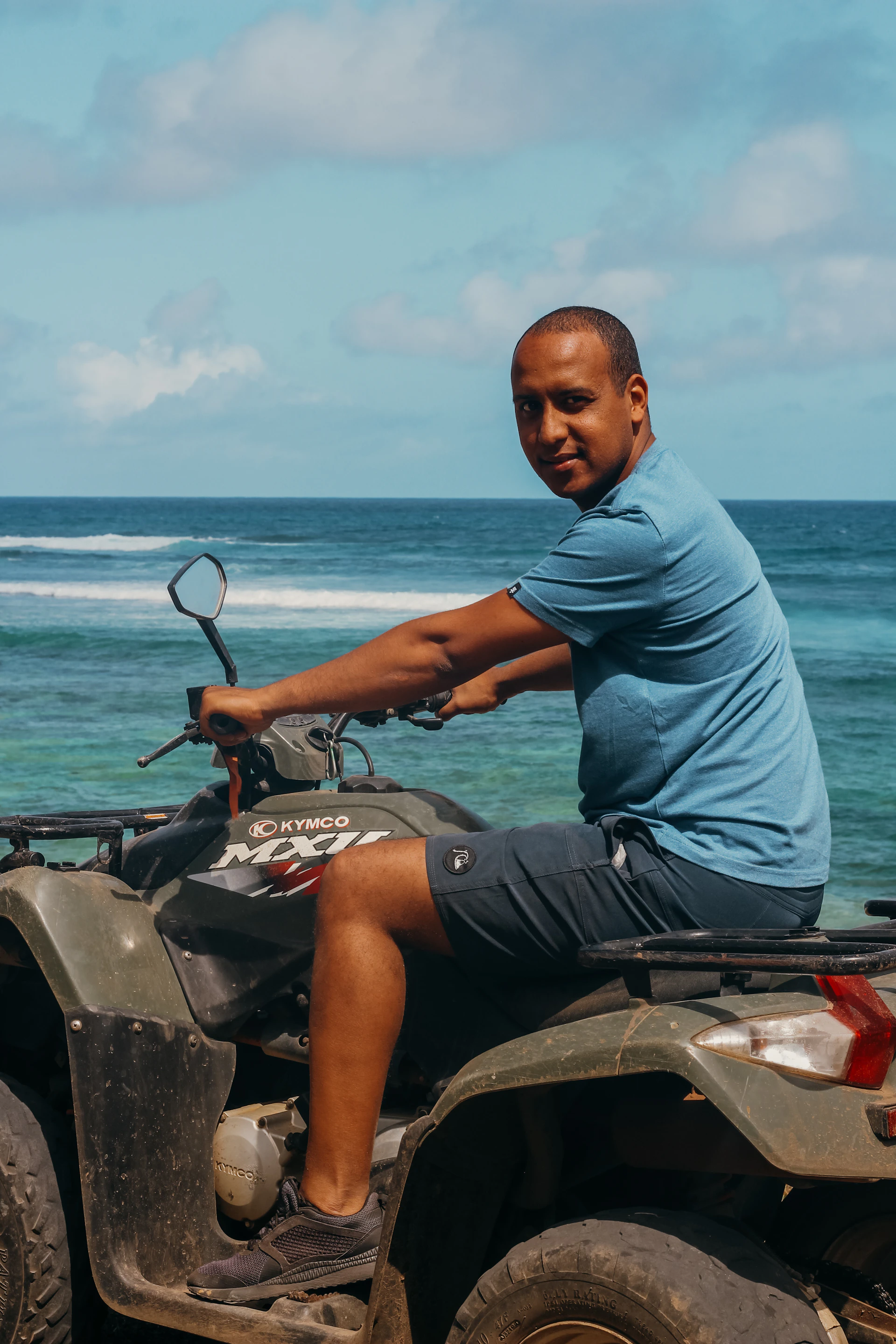 Close-up of a rider gripping the handlebars of an ATV, with the sparkling ocean and clear blue sky behind.