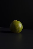 A single lime is placed on a dark surface with a black, textured background. The lighting casts a soft shadow, highlighting the lime's textured, green skin and slightly uneven shape.