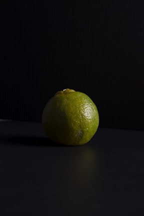 A single lime is placed on a dark surface with a black, textured background. The lighting casts a soft shadow, highlighting the lime's textured, green skin and slightly uneven shape.