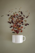 A white ceramic mug is placed on a soft green surface, with a variety of loose tea leaves, dried fruits, and herbs artistically scattered above it, as if they are being poured into the mug.