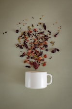 A white ceramic mug is placed on a soft green surface, with a variety of loose tea leaves, dried fruits, and herbs artistically scattered above it, as if they are being poured into the mug.