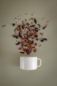 A white ceramic mug is placed on a soft green surface, with a variety of loose tea leaves, dried fruits, and herbs artistically scattered above it, as if they are being poured into the mug.