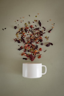 A white ceramic mug is placed on a soft green surface, with a variety of loose tea leaves, dried fruits, and herbs artistically scattered above it, as if they are being poured into the mug.