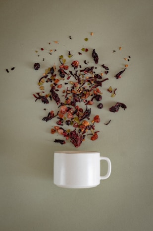A white ceramic mug is placed on a soft green surface, with a variety of loose tea leaves, dried fruits, and herbs artistically scattered above it, as if they are being poured into the mug.
