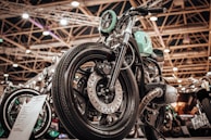 A close-up view of a custom motorcycle with a vintage design featuring a mint-green fuel tank and chrome detailing. The setting is an indoor exhibition hall with bright overhead lighting and structural beams visible in the background.