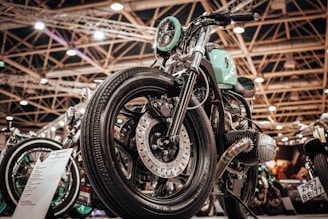 A close-up view of a custom motorcycle with a vintage design featuring a mint-green fuel tank and chrome detailing. The setting is an indoor exhibition hall with bright overhead lighting and structural beams visible in the background.