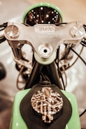 A close-up view of a motorcycle's handlebars and top components, featuring a silver metallic handlebar assembly with visible bolts and wiring. The brand name BMW is prominently engraved on the central piece. The foreground shows a green and black tank with intricate metallic detailing on top.