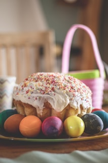 A round cake with white frosting and multicolored sprinkles is placed on a plate surrounded by vividly colored Easter eggs. The background includes a softly focused pink basket and a wooden chair, contributing to a casual, festive atmosphere.