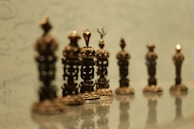A row of ornate, intricately designed metallic chess pieces lined up in a line, with a soft greenish background. The pieces are highly detailed with a classical design, potentially handcrafted, and the focus softens as it moves towards the background.