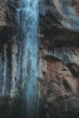 A person is rappelling down a waterfall situated on a rugged cliffside. The cascading water flows dramatically over the rocks, creating a dynamic scene. The climber is secured with gear and rope, appearing focused and adventurous.