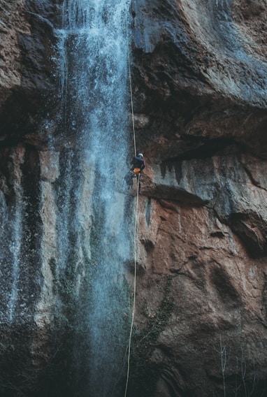 A person is rappelling down a waterfall situated on a rugged cliffside. The cascading water flows dramatically over the rocks, creating a dynamic scene. The climber is secured with gear and rope, appearing focused and adventurous.