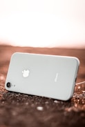 A smartphone with a white back and silver apple logo is lying on a rough surface, angled with the camera lens visible in the lower part of the image. The lighting gives a warm glow to the background, suggesting a sunset or sunrise.