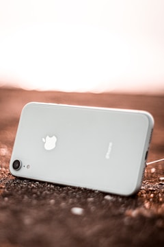 A smartphone with a white back and silver apple logo is lying on a rough surface, angled with the camera lens visible in the lower part of the image. The lighting gives a warm glow to the background, suggesting a sunset or sunrise.