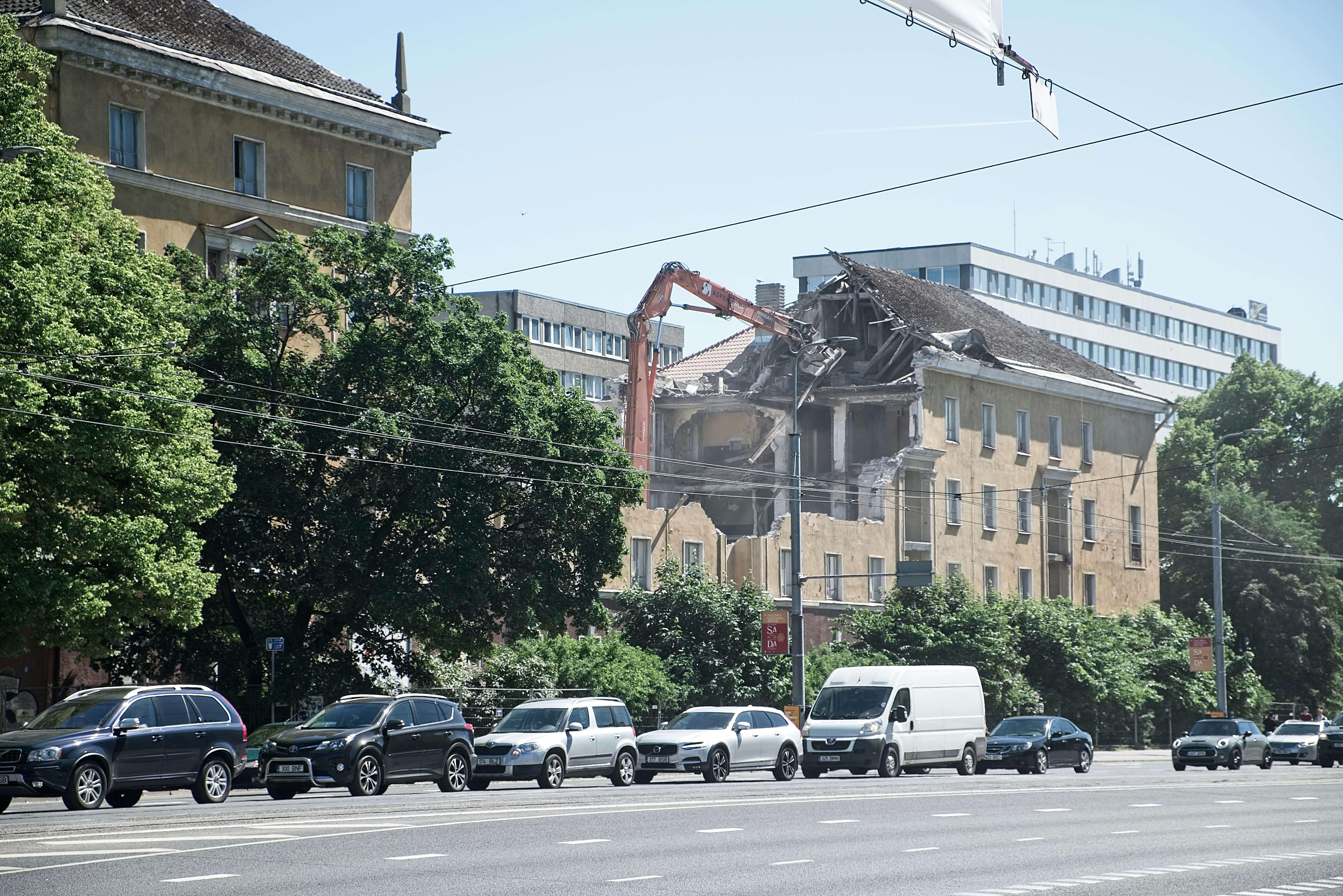 A partially demolished building with a construction machine actively tearing down the structure while cars pass by on the street. Green trees line the street, contrasting with the urban scene. The sky is clear and blue.
