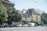 A partially demolished building with a construction machine actively tearing down the structure while cars pass by on the street. Green trees line the street, contrasting with the urban scene. The sky is clear and blue.