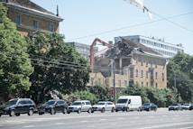 A partially demolished building with a construction machine actively tearing down the structure while cars pass by on the street. Green trees line the street, contrasting with the urban scene. The sky is clear and blue.