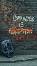 Graffiti on a brick wall reads 'Happiness is a perception chalice' with colorful paint. A child’s car seat is positioned on the pavement in front of the wall.