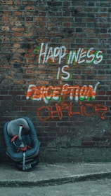 Graffiti on a brick wall reads 'Happiness is a perception chalice' with colorful paint. A child’s car seat is positioned on the pavement in front of the wall.