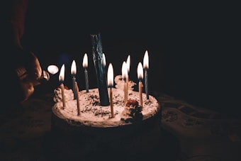 A cake topped with multiple lit candles, surrounded by a dark background. A hand is holding a match, likely to light more candles. The cake is decorated with swirls of cream and sprinkles. The image is dimly lit, with the candles providing the main source of light.