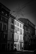 A street scene featuring a row of classic European buildings with shuttered windows and wrought iron balconies. The lighting highlights the contrast between shadow and illumination, creating dramatic tension. Above the street, a star-shaped light fixture is suspended, suggesting a festive or holiday theme. A cyclist is captured in motion at the bottom center of the image, adding an element of urban life.