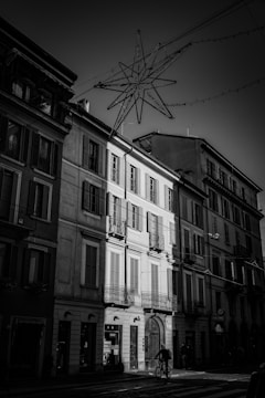A street scene featuring a row of classic European buildings with shuttered windows and wrought iron balconies. The lighting highlights the contrast between shadow and illumination, creating dramatic tension. Above the street, a star-shaped light fixture is suspended, suggesting a festive or holiday theme. A cyclist is captured in motion at the bottom center of the image, adding an element of urban life.