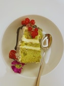 A slice of layered green tea cake with cream filling, garnished with red currants and a decorative purple flower, is placed on a white plate. A silver spoon lies beside the cake.