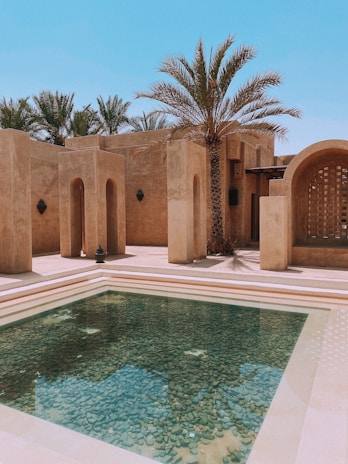 A serene outdoor scene features a luxurious pool surrounded by stone architecture with tall arches and textured walls. Palm trees rise in the background under a clear blue sky, adding a tropical touch to the tranquil setting.