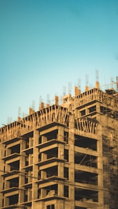 A partially constructed building with concrete floors and vertical beams extending from the top. The structure is exposed without walls, and the surrounding area is clear blue sky, suggesting an unfinished skyscraper or a high-rise under construction.