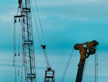 Two large industrial structures are set against a blue sky. One structure appears to be a red crane with several pulleys and cables, and the other is a large yellow mechanical arm, possibly part of construction or oil extraction equipment.