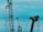 Two large industrial structures are set against a blue sky. One structure appears to be a red crane with several pulleys and cables, and the other is a large yellow mechanical arm, possibly part of construction or oil extraction equipment.