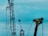 Two large industrial structures are set against a blue sky. One structure appears to be a red crane with several pulleys and cables, and the other is a large yellow mechanical arm, possibly part of construction or oil extraction equipment.