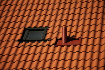Red clay roofing tiles cover a sloped surface. A small, black-framed skylight is situated on the left side, while a red vent pipe protrudes from the right, both integrated into the tiled pattern.