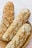 Three rustic loaves of bread with a golden, crusty exterior rest in a woven basket. The bread surface is uneven with a textured crust, suggesting a hearty, artisanal quality.