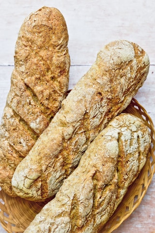 Three rustic loaves of bread with a golden, crusty exterior rest in a woven basket. The bread surface is uneven with a textured crust, suggesting a hearty, artisanal quality.