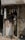 A small, dimly lit shop with an elderly man inspecting items on a cluttered counter. The shop has a faded, weathered exterior with a metal rolling shutter partly closed. Various small products hang from the ceiling, and there is a poster on the front of the counter.