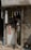 A small, dimly lit shop with an elderly man inspecting items on a cluttered counter. The shop has a faded, weathered exterior with a metal rolling shutter partly closed. Various small products hang from the ceiling, and there is a poster on the front of the counter.