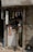 A small, dimly lit shop with an elderly man inspecting items on a cluttered counter. The shop has a faded, weathered exterior with a metal rolling shutter partly closed. Various small products hang from the ceiling, and there is a poster on the front of the counter.