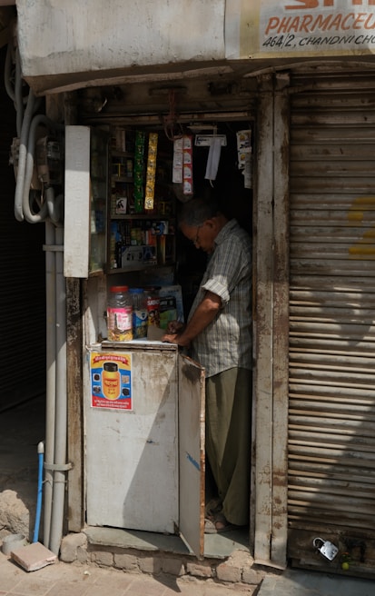 A small, dimly lit shop with an elderly man inspecting items on a cluttered counter. The shop has a faded, weathered exterior with a metal rolling shutter partly closed. Various small products hang from the ceiling, and there is a poster on the front of the counter.