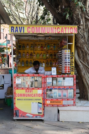 A small kiosk under a large tree, selling mobile accessories and repair services. It is named 'Ravi Communication', as indicated by signs on the storefront. The stand is neatly arranged with mobile phone covers and various accessories. A person is seated inside, seemingly attending to tasks.