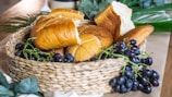 A woven basket is filled with a variety of freshly baked bread and clusters of dark grapes. The assortment includes different types of bread with golden brown crusts. Surrounding the basket are green leaves and a wooden surface that adds to the rustic setting.