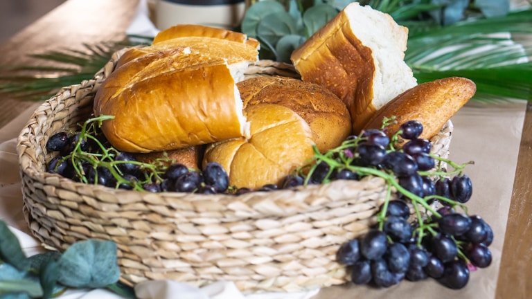 A woven basket is filled with a variety of freshly baked bread and clusters of dark grapes. The assortment includes different types of bread with golden brown crusts. Surrounding the basket are green leaves and a wooden surface that adds to the rustic setting.