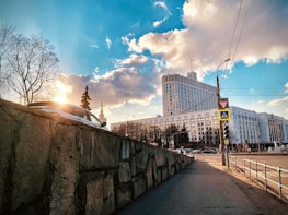 A cityscape featuring a large government or office building with a modern architectural design. The structure is surrounded by bare trees on a clear day with few clouds. A car is parked on a raised stone wall to the left, while traffic signs and street lamps are visible along the sidewalk.
