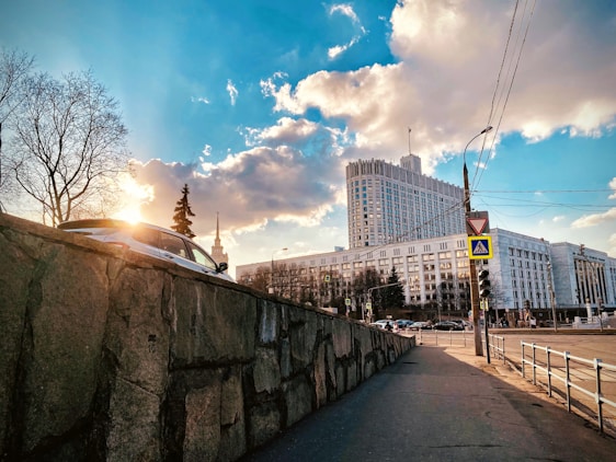 A cityscape featuring a large government or office building with a modern architectural design. The structure is surrounded by bare trees on a clear day with few clouds. A car is parked on a raised stone wall to the left, while traffic signs and street lamps are visible along the sidewalk.