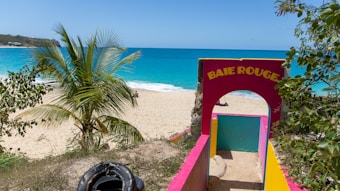 A tropical beach scene featuring a vibrant entryway labeled 'Baie Rouge' in bold yellow letters against a bright pink arch. Lush green palm trees frame the sandy beach, which borders the vivid turquoise sea under a clear blue sky.