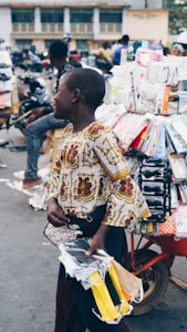 A person wearing a patterned shirt stands on a busy street near a display of various packaged items hanging from a cart. The individual is holding a collection of colorful fabric masks. There are motorcycles and other people visible in the background, suggesting a bustling marketplace setting.
